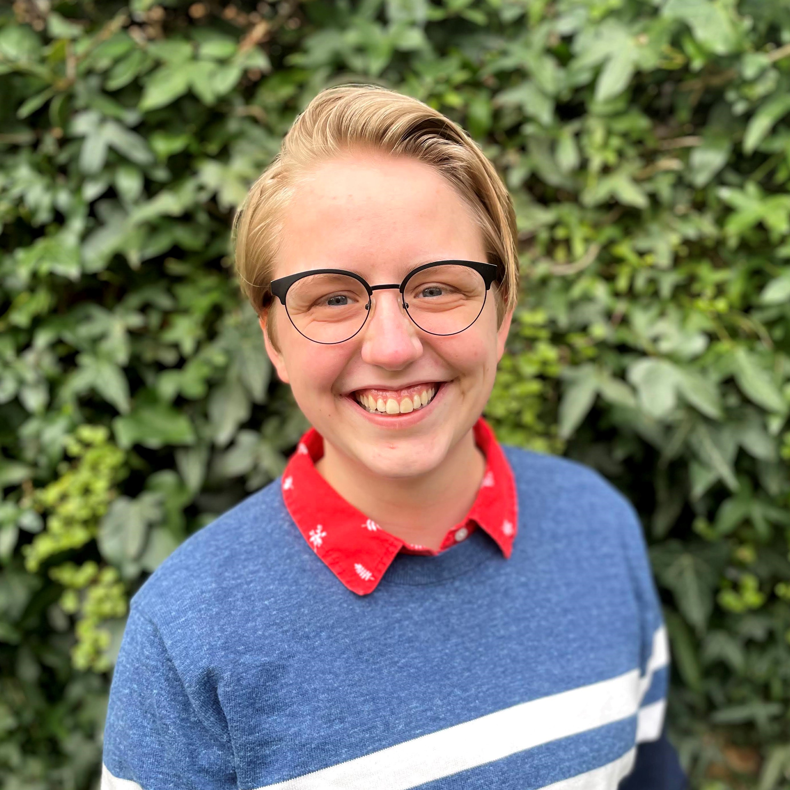 Claire, a white non-binary person with short blond hair wearing glasses, smiles at the camera in front of a wall covered in leaves.