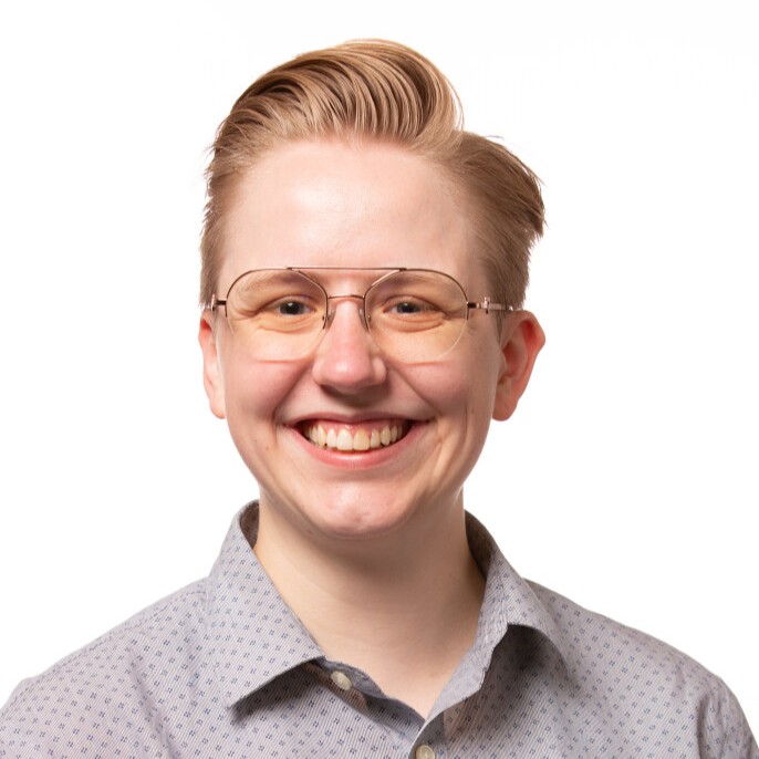 Claire, a white non-binary person with short blond hair wearing glasses, smiles at the camera in front a white background.